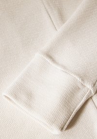 Nahaufnahme eines hellbeigen, strukturierten Stoffes mit einem weichen, erhabenen Muster und einem gefalteten Umschlag mit sauberen Nähten entlang der Kanten.