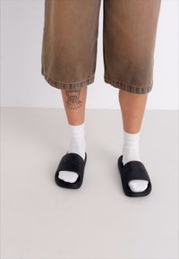 Sorte slidesandaler med forhøjet logo, båret med hvide ankelstrømper, parret med brune knælange shorts. Synlig tatovering på benet.