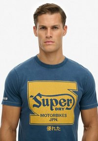 Jonge man draagt een blauw T-shirt met een geel "Super Dry Motorbikes JPN" logo en Japanse tekens op een effen witte achtergrond.