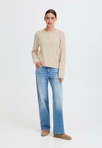 Beige neulecardigan, jossa on teksturoitu kuvio, pyöreä pääntie ja nappikiinnitys, yhdistettynä vaaleansiniset suorat farkut ja beigeet mokka-nahkasaappaat.