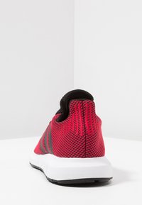 Röd och svart träningsskor med ett meshtyg på ovansidan, texturerad baksida och vit sula med svarta accenter och tre svarta ränder.