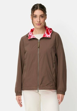 Frau trägt eine braune Jacke mit Reißverschluss und eine pink-rote gemusterte Kapuze, kombiniert mit hellen Hosen, und steht vor einem schlichten Hintergrund.