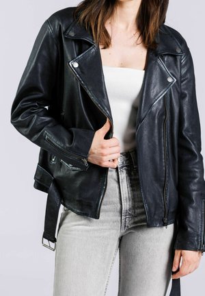 Persona que lleva una chaqueta de cuero negra, top blanco acanalado y vaqueros de talle alto gris claro, sujetando la cremallera de la chaqueta con una mano.