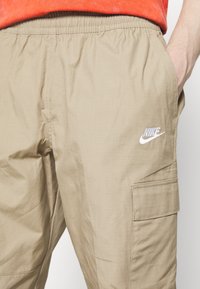 Beige cargobyxor med elastisk midja, sidfickor och en broderad vit Nike-logotyp, tillverkade av mjukt och lätt tyg.