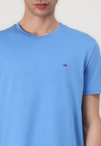 Blaues kurzärmeliges T-Shirt mit Rundhalsausschnitt aus Baumwollstoff, mit einem kleinen bestickten Logo in Rot, Weiß und Blau auf der linken Brust.