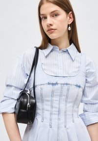 Νεαρή γυναίκα με ίσια καστανά μαλλιά, που φοράει ανοιχτό μπλε ριγέ πουκάμισο και ξεβαμμένο τζιν φόρεμα, κρατώντας μαύρη τσάντα ώμου, με εμφανές σκουλαρίκι με μαργαριτάρι.