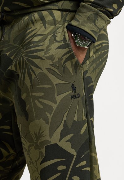 Olivově zelené kalhoty se vzorem velkých listů, vyrobené z měkké látky. Obsahují viditelné logo a kapsu, z níž je částečně vidět hodinky.