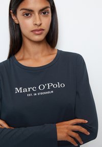 Granatowa koszula z długim rękawem od Marc O'Polo z białym nadrukowanym logo i tekstem. Miękka tkanina z okrągłym dekoltem, zaprezentowana na modelu.