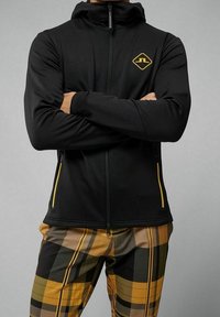 Felpa nera con zip realizzata in tessuto elasticizzato, con un logo giallo sul petto e dettagli gialli sulla zip. Presenta un design aderente e un colletto alto.