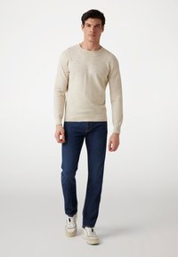 Beige Langarmpullover mit strukturiertem Finish, kombiniert mit blauen Jeans und weißen Sneakers, vor einem einfarbigen Hintergrund stehend.