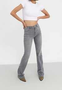 Harmaat hulkkareiden farkut, joissa on sileä pinta, yhdistetty valkoiseen istuvaan lyhytvartiseen t-paitaan. Seisova poseeraus, kädet lantiolla.