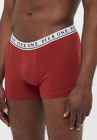 Rote Baumwoll-Boxershorts mit einem schwarz-weißen, markierten Bund, die eine enge Passform und eine glatte Textur bieten.