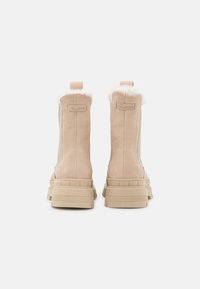 Beige suede ankle boots with thick rubber soles and white fur trim, shown from the back on a white background.