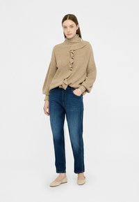 Maglione beige in maglia con dettagli a volant, collo alto, maniche lunghe. Abbinato a jeans blu in denim e scarpe basse di colore chiaro. Sfondo bianco.