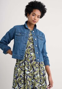 Veste en jean de couleur bleu moyen, style court avec fermeture à boutons, col rabattu et deux poches poitrine, portée par-dessus une robe à motifs floraux.