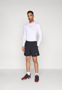 Άσπρο πουκάμισο με μακριά μανίκια και μαύρο λογότυπο, μαύρο αθλητικό σορτς και μαύρα/κόκκινα αθλητικά παπούτσια. Το μοντέλο στέκεται σε ουδέτερο φόντο.