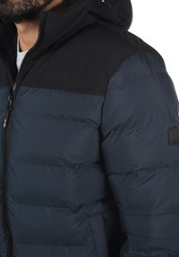 Tamno plavi puffer jakna s crnim ramenskim panelom, s patentnim zatvaračem, kapuljačom i teksturiranim materijalom sa suptilnim sjajem.