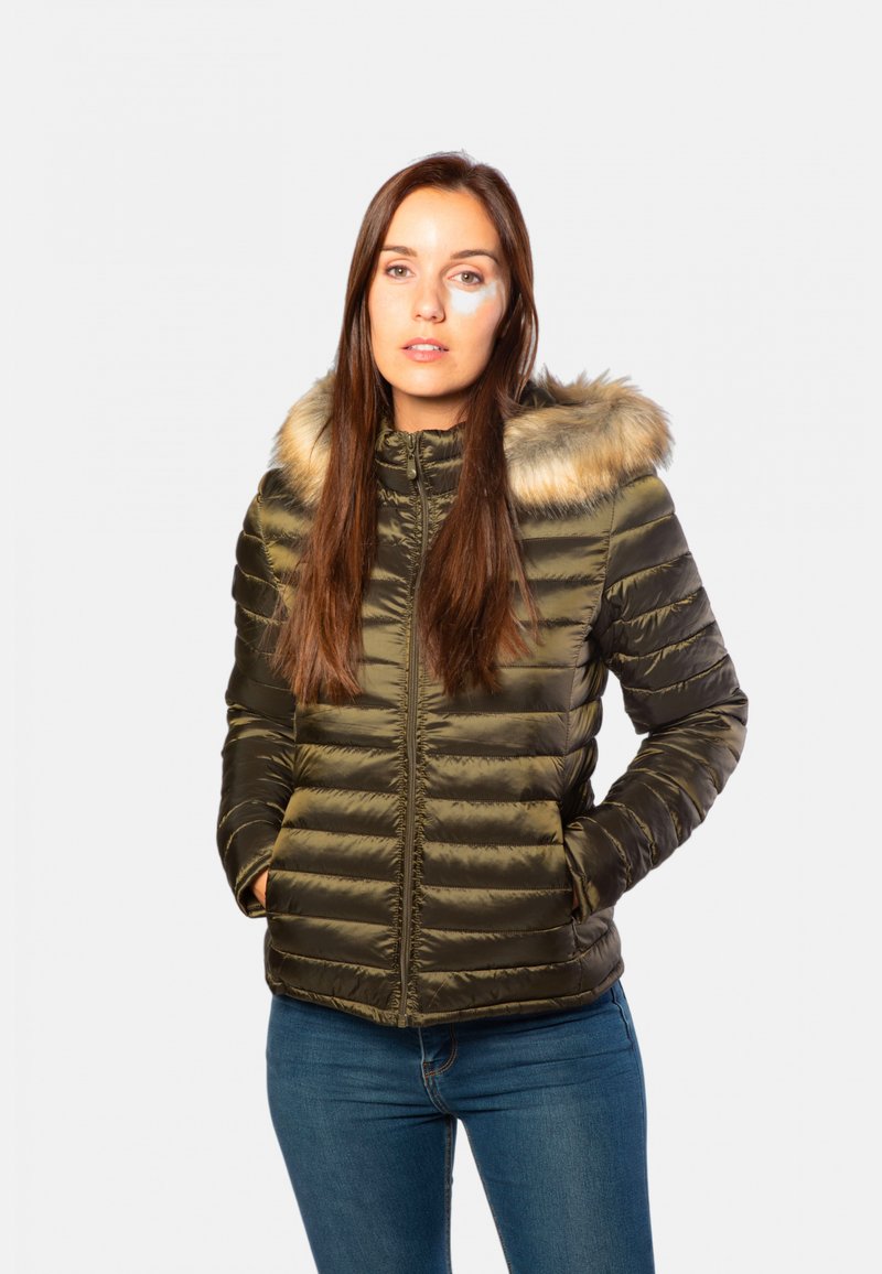 Veste matelassée vert olive avec une bordure en fausse fourrure sur la capuche, fermeture éclair et motif matelassé horizontal. Portée avec un jean bleu.