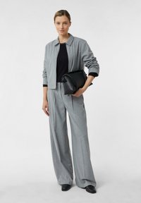 Grauer Stoffanzug mit einem kragenbesetzten Blazer und weit geschnittenen Hosen. Schwarzes Oberteil darunter. Das Model hält eine schwarze Handtasche. Glatte Textur und neutrale Farbtöne.