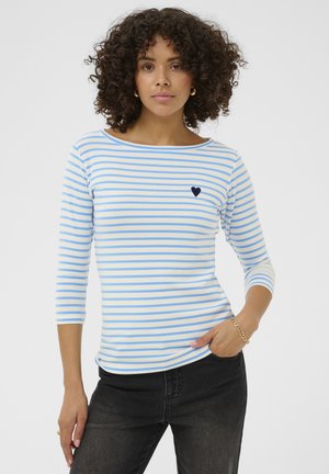 Donna con capelli ricci che indossa una camicia a maniche lunghe bianca e blu a righe con un piccolo cuore blu scuro, pantaloni neri e un braccialetto d'oro.