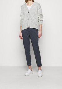 Grå, stickad kofta med V-ringning, fem svarta knappar och ribbade ärmslut, matchad med mörka byxor och vita sneakers.