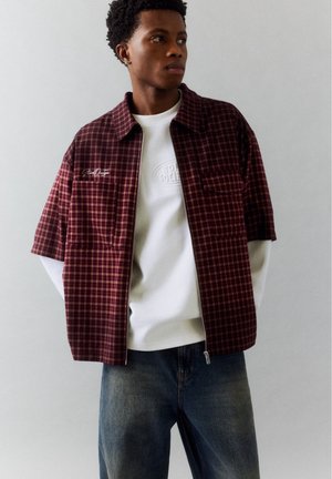 Un homme porte une chemise à manches courtes à carreaux bordeaux et à fermeture éclair par-dessus un haut blanc et un jean foncé, devant un fond clair uni.