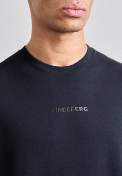 T-shirt en coton noir avec un col rond, présentant un logo texturé "J.LINDEBERG" sur la poitrine, tissu lisse et manches courtes.