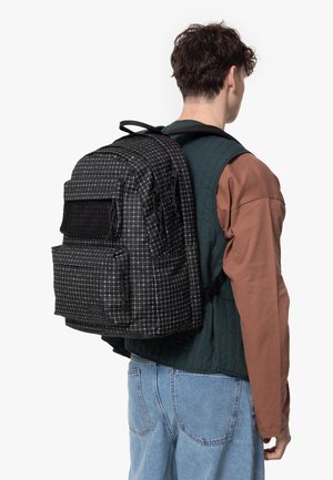 Black and white checkered backpack with multiple pockets, a mesh front pocket, and padded straps. Worn on a person, features quilted vest.