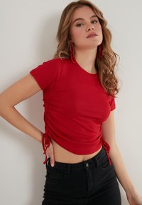 Červený krátký rukáv top s řasením na bocích a zavazováním, doplněný o černé džíny s vysokým pasem. Modelka má na sobě červené náušnice.