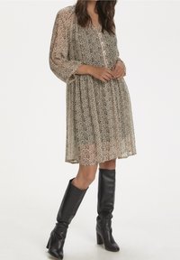 Robe beige transparente avec un motif abstrait noir, dotée de manches longues, d'une coupe ample et d'une longueur mi-mollet, assortie à des bottes noires montant jusqu'au genou.