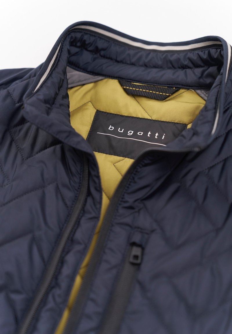 Marineblaue gesteppte Jacke mit gelb gefüttertem Innenfutter, ausgestattet mit einem Reißverschluss und einem Etikett mit der Aufschrift "bugatti" im Kragen.