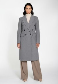 Grå dobbeltradet frakke i struktureret stof med sorte knapper og sidelommer. Båret over en beige turtleneck og brede beige bukser.