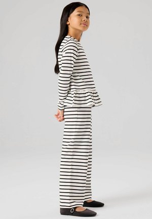 Ragazza giovane in piedi con maglia a righe a maniche lunghe con volant e pantaloni larghi abbinati, indossa scarpe nere Mary Jane, sfondo neutro.