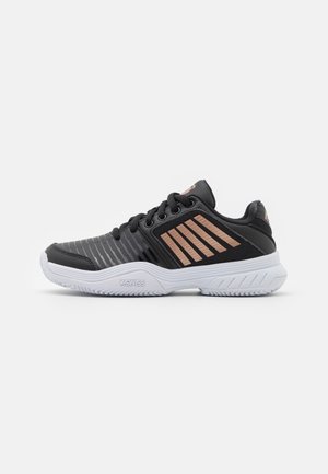 Chaussure de sport noire avec des rayures grises, des accents en or rose et une semelle blanche. Comprend des lacets et des surfaces texturées. Marque KSwiss sur le côté.