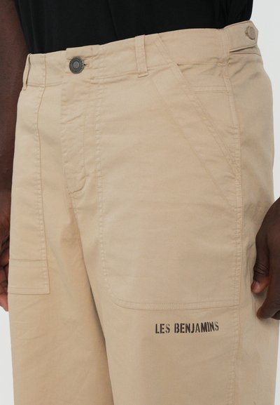 Pantalons cargo beige en coton, avec poches latérales, fermeture à bouton et le texte "LES BENJAMINS" imprimé en noir à l'arrière.