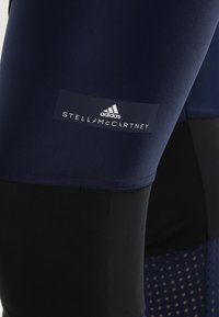 Navy urheiluleggingsit mustilla yksityiskohdilla, jossa on sileä pinta, logo-paneeli valkoisella tekstillä ja hyvin tuuletettavat verkko-osat.