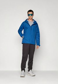 Modra nepremočljiva jakna s kapuco, siva majica, temne hlače in svetli supergi. Model nosi odsevna očala za sonce.