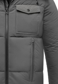 Grau gepolsterte Jacke mit Reißverschluss, einer Brusttasche mit Klappe und einer vertikalen Seitentasche mit Reißverschluss. Glatte Stoffoberfläche.