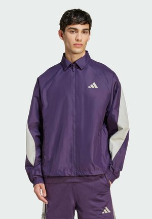 Veste légère violette avec des accents gris, fermeture éclair intégrale, poignets côtelés et un logo sur la poitrine gauche. Convient pour les vêtements de sport.