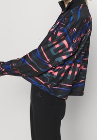 Osoba v černé bundě s abstraktními růžovými a modrými vzory, zkombinovaná s černými legínami Adidas, stojící bokem na neutrálním pozadí.