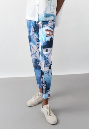 Osoba ubrana w niebieskie spodnie z nadrukiem nawigacyjnym przedstawiającym żaglówki, mewy i kompas, w białych trampkach, stojąca na szarej podłodze.