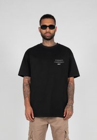Black cotton t-shirt with short sleeves, featuring white text and graphics on the chest. Worn with beige cargo pants and sunglasses.