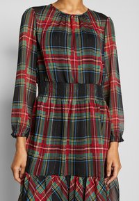 Robe à manches longues avec un motif à carreaux rouge, vert, bleu et jaune, taille froncée et poignets élastiques sur tissu transparent.