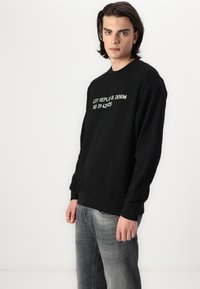 Sudadera negra con texto impreso en blanco en la parte frontal, con cuello redondo y puños acanalados. Combinada con unos vaqueros de denim gris claro.