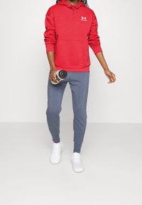Roter Kapuzenpullover mit Fronttasche und weißem Logo, kombiniert mit blauen strukturierten Jogginghosen und weißen Sportschuhen, hält eine Wasserflasche.