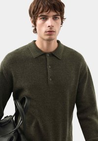 Ciemnozielony sweter z dzianiny z kołnierzykiem, z zapięciem na guziki i teksturowanym materiałem. Pod pachą trzymana jest czarna torba.