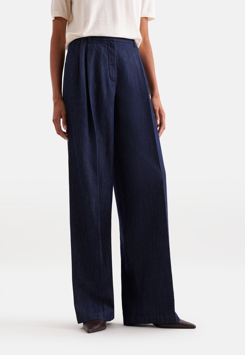 Femme portant un pantalon en jean bleu foncé à jambes larges et des chaussures marron pointues, associé à un top blanc à manches courtes.
