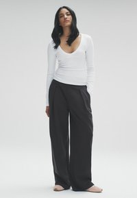 Top bianco a maniche lunghe con scollatura a V e design aderente abbinato a pantaloni scuri a gamba larga. Il modello indossa scarpe slip-on di colore chiaro.