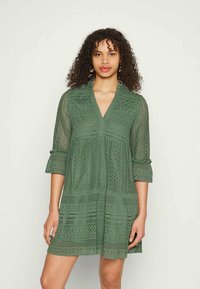Vestido verde renda com decote em V, mangas três quartos e padrões intrincados. O tecido tem um acabamento texturizado e um corte descontraído.