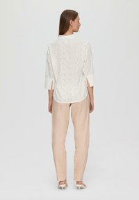 White shirt with eyelet detailing, three-quarter sleeves, and a rounded hem, paired with light beige linen pants and light-colored footwear.